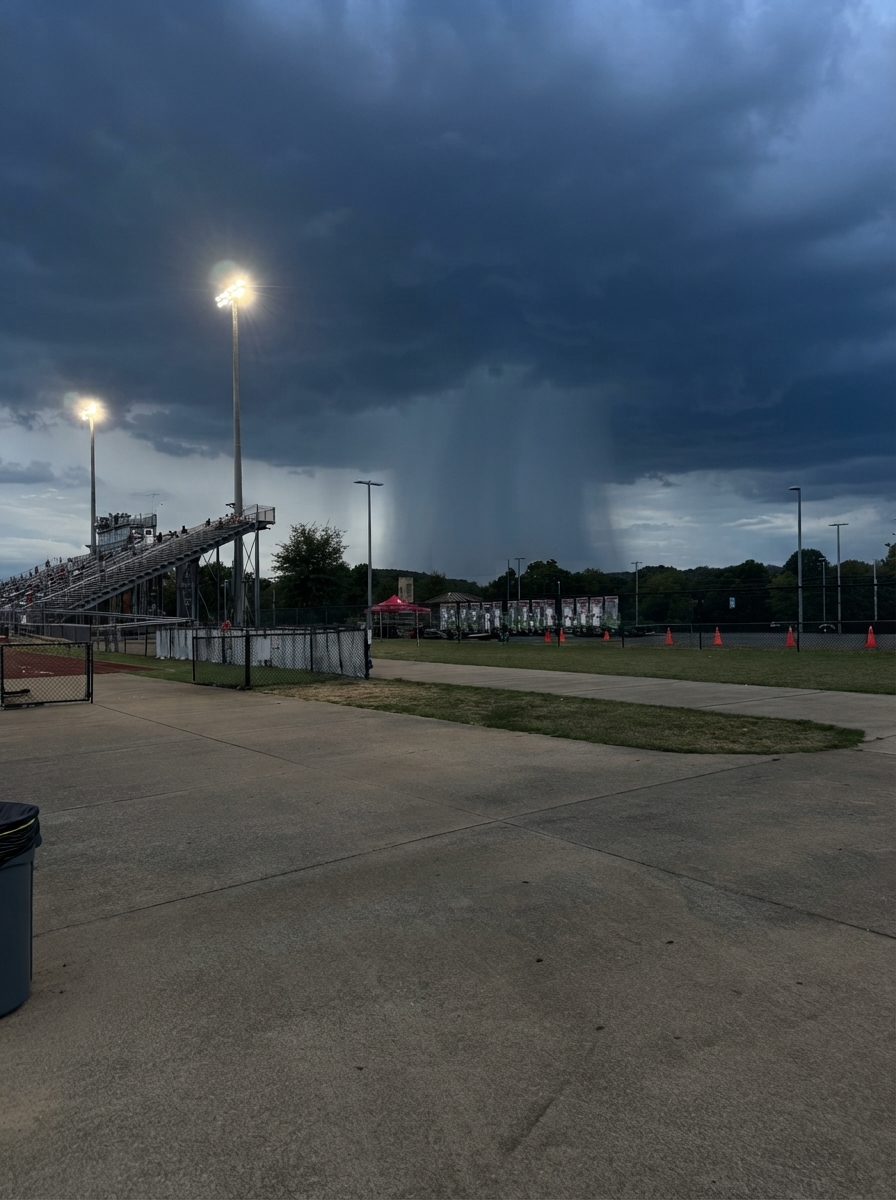 High-school stadium ten minutes later, empty of spectators, with the rain column still visible behind the tree line