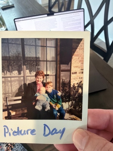 A hand holding an old instant photo labeled Picture Day: a woman on a porch bench with two young children