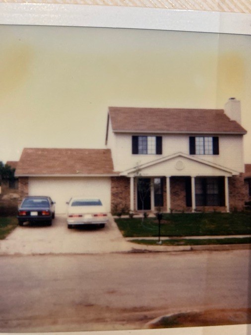A faded 1980s print of a two-story brick and siding house with two cars in the driveway