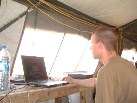 A young soldier working at a laptop on a plywood desk inside a tent