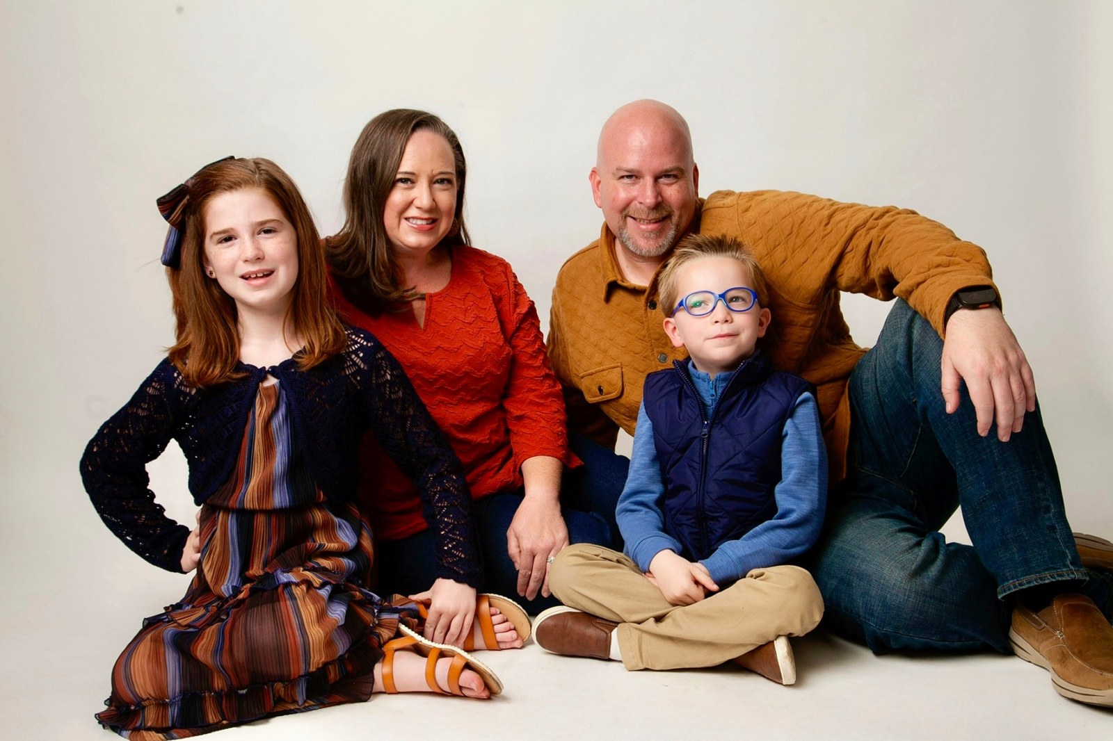 A studio family portrait: a mother, a father, a daughter and a young son in glasses, seated together on a white floor